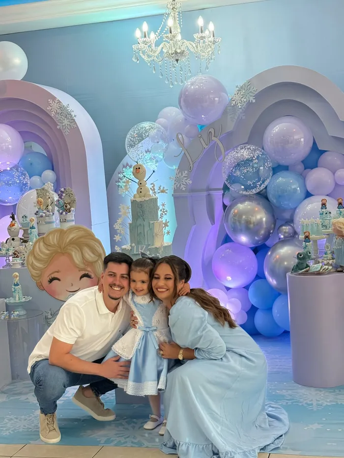 Família posando com a filha diante do cenário com tema Frozen, arcos de balões e bolo com o Olaf